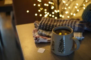A beige ceramic mug featuring an etched pine tree design sits on a wooden table, filled with tea with its tea bag string hanging over the side. In the soft-focused background, folded patterned textiles are stacked next to the mug beneath warm, glowing fairy lights.