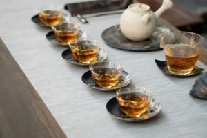 A Gongfu tea setup is arranged on a light grey runner, featuring a ceramic side-handled teapot and a large glass pitcher filled with amber tea. Five small, scalloped glass teacups filled with the same amber liquid are lined up diagonally on dark, patterned saucers.