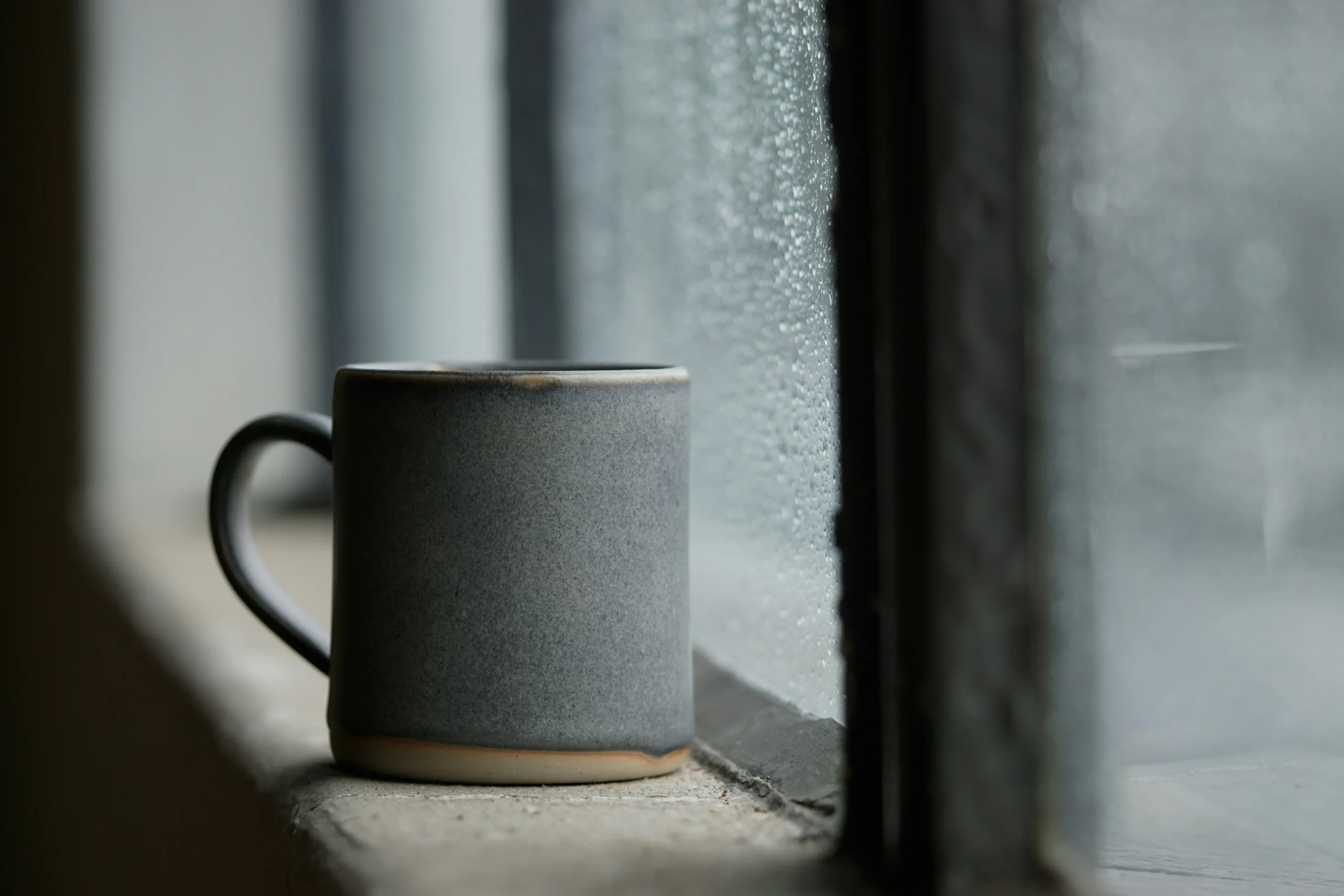 Why Tea Somehow Tastes Better on Rainy Days A matte grey ceramic mug sits on a concrete windowsill next to a window covered in raindrops. The background shows a soft, out-of-focus view of a gloomy, rainy day.