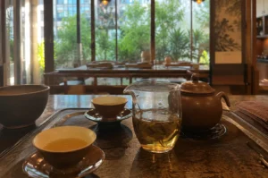 A serene and traditional tea service setup is arranged on a dark, textured tray over a polished wooden counter. In the foreground, the service features a clear glass pitcher (fairness cup) filled with amber-colored tea and visible loose tea leaves, a round clay teapot sitting on a small trivet, and two small ceramic teacups resting on matching saucers, one of which is filled with tea. To the far left, a larger dark ceramic bowl sits just off the tray. The background opens up to a warm, sunlit tea house interior, revealing wooden tables and chairs set for guests, warm hanging pendant lights, and a large floor-to-ceiling glass window that looks out onto a lush courtyard filled with vibrant green trees and plants.