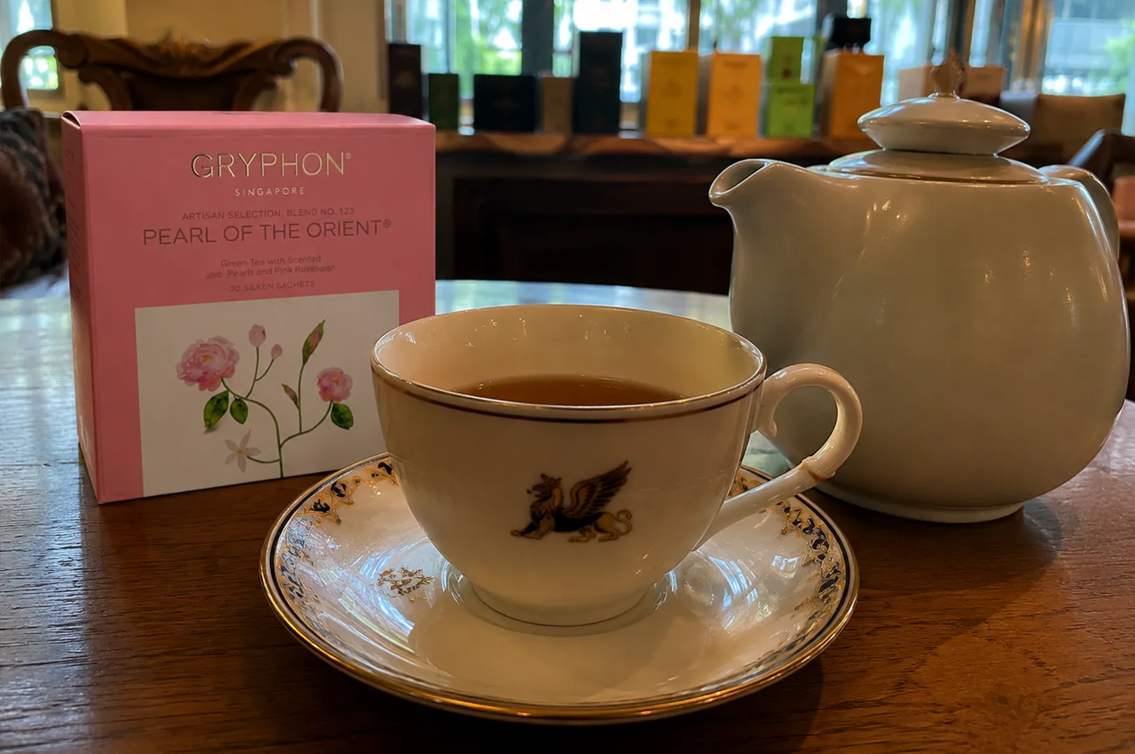 1 - Discovering Gryphon Tea Company’s Earl Grey in Singapore A refined afternoon tea setup is elegantly arranged on a polished wooden table. In the center foreground sits a delicate white porcelain teacup filled with clear amber tea, resting on a matching saucer featuring an ornate navy blue and gold patterned rim with a gilded edge; the front of the teacup is emblazoned with a prominent gold griffin emblem. To the left of the cup, a square, pastel pink tea box from "Gryphon Singapore" stands upright, clearly labeled "Pearl of the Orient" with an illustration of flowering pink roses on its lower half. To the right, a large, rounded ceramic teapot in a soft, pale celadon green completes the tea service. The blurred background features a warm interior with a classic wooden chair on the left and a row of vibrantly colored, assorted tea boxes neatly displayed along a shelf in front of large windows that let in soft light and offer a glimpse of green trees outside.