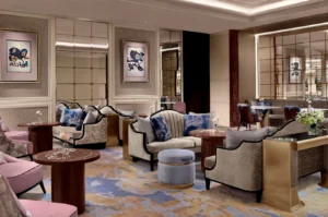 This wide shot captures an elegant and luxurious hotel lounge decorated in sophisticated muted tones, featuring plush seating arrangements spaced across a large, patterned carpet with swirling shades of blue, gold, and cream. The main seating area includes curved beige sofas adorned with deep blue and silver-patterned throw pillows, paired with small round wooden coffee tables, a low grey velvet ottoman with a gold base, and armchairs with soft pink velvet upholstery. The walls are finished in a neutral textured wallpaper, punctuated by large grid-patterned mirrors that reflect the room and modern abstract framed artwork, creating an inviting yet upscale ambiance.