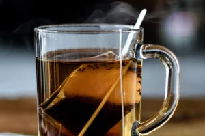 A close-up side view captures a clear glass mug filled with hot, dark tea, with a tea bag still steeping inside. A white tag on a string hangs over the rim of the mug, and delicate wisps of steam rise from the surface of the tea, indicating that the drink is freshly brewed and hot.