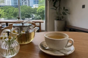 A serene cafe scene showcases a speckled, off-white ceramic teacup filled with light yellow tea resting on a saucer with a silver spoon, positioned on a smooth wooden table. To the left sits a clear glass teapot with a stainless steel infuser brewing tea, next to a small, ribbed glass vase holding fresh chamomile flowers. In the background, large glass windows reveal a lush view of green trees and city buildings, while a potted indoor plant stands in the softly lit, minimalist cafe interior.