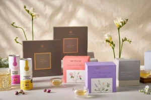 A balanced product display showcases an assortment of Gryphon Tea items arranged on a light gray surface against a textured beige fabric backdrop. In the center and foreground are several cube-shaped artisan tea boxes in pastel pink (Pearl of the Orient), lavender (Earl Grey Lavender), and light gray (Earl Grey), accompanied by two taller, dark brown "Monogram" gift boxes standing behind them. To the left, two aluminum tea cans—one white and pink (Mogambo) and one white and yellow (Osmanthus Sencha Passionfruit)—sit next to a tall glass filled with iced tea, while two small, double-walled glass cups containing warm, amber-colored tea are placed out front. The serene, elegant setup is completed by delicate white flowers on long green stems framing the arrangement on both sides.