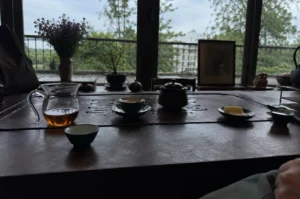 A moody, low-lit indoor tea-drinking setup is arranged on a large, textured dark brown wooden table, facing a wall of large glass windows that look out onto a rainy or overcast day. In the foreground, several small, dark-colored ceramic teacups and saucers are scattered across the table, along with a small, lidded dark teapot and a small plate holding a light-colored snack or pastry. To the left, an amber-colored liquid fills a clear glass pitcher. Outside the windows, a balcony railing overlooks a lush green landscape of trees, with a building visible in the distant background under a cloudy grey sky. On the windowsill or balcony area, decorative items such as a vase of purple flowers and a small potted plant add life to the serene, contemplative atmosphere.
