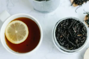 A bright, minimalist scene showing a white cup of amber-colored tea with a slice of lemon floating on top, placed next to a glass jar filled with dark Lapsang Souchong tea leaves, set against a clean, marble-patterned countertop.
