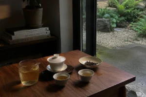A serene, high-angle shot depicts a contemplative tea ritual set on a dark, polished wooden table beside an open doorway looking out onto a lush, rain-kissed Japanese garden. On the table, a delicate white gaiwan sits on a dark ceramic tray, accompanied by two small, filled teacups and a glass fair cup containing golden, steeped tea, while a shallow bowl to the side holds loose, dark tea leaves. In the soft, dim light, a stack of minimalist hardback books—titled "This is Home," "Kinfolk," and "The Touch"—sits on a shelf in the background beneath a small potted plant, enhancing the quiet, reflective atmosphere of the scene.