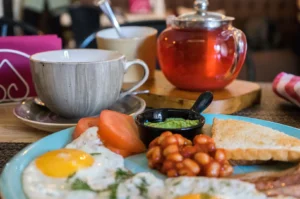 A vibrant breakfast spread is presented on a table, with the foreground focusing on a turquoise plate holding two fried eggs garnished with dill, sliced fresh tomatoes, baked beans in tomato sauce, a small black dish of mashed avocado, and a piece of toast. In the soft-focused background, an elegant gray textured teacup sits on its saucer next to a clear glass teapot filled with reddish-amber tea resting on a wooden board.