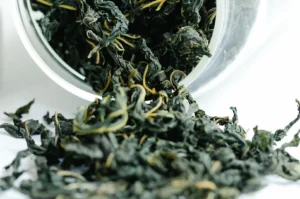 This macro shot shows a glass jar tipped onto its side, with dry, dark, tightly rolled Tie Guan Yin tea leaves spilling out onto a clean white surface. The leaves display their characteristic deep green and brownish hues, with lighter stems visible, emphasizing the quality and raw texture of the premium Oolong tea.