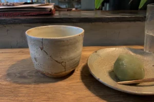 A close-up, eye-level shot features a striking example of kintsugi art on a wooden counter. On the left stands a light beige ceramic cup with a rustic, unglazed base, displaying prominent cracks beautifully mended with dark gold lacquer. To the right, a shallow, textured grey ceramic plate holds a single, round green mochi sweet with a small wooden pick resting against it. The background is dimly lit and out of focus, showing a dark stone counter lip and glimpses of papers and green leaves, keeping the emphasis on the repaired pottery.