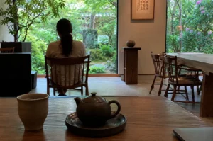A serene and contemplative scene unfolds within a minimalist, Zen-inspired tea space, captured from a low perspective at a foreground wooden table. Resting on the table in the immediate foreground is a rustic, light brown ceramic teacup and a small, matte dark brown teapot sitting on a matching circular saucer, alongside the edge of a closed laptop on the bottom right. In the midground, a woman with dark hair tied back in a ponytail sits with her back to the camera in a wooden chair, quietly gazing out through a massive, floor-to-ceiling glass window. The window frames a beautiful view of a lush, vibrant green Japanese-style garden complete with thick foliage, ferns, and a moss-covered stone lantern. To the right of the window, a clean cream-colored wall is decorated with a framed piece of elegant Chinese or Japanese calligraphy, beneath which a tall, narrow wooden stand supports a small decorative bowl, while further to the right, a long wooden communal table with matching wooden chairs sits near a window showcasing blooming purple flowers.