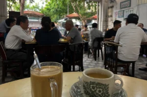 A round table holds a tall, clear glass mug filled with a light brown, milk-coffee beverage with a spoon resting inside, alongside a traditional ceramic teacup and saucer adorned with a green floral pattern, filled with a dark tea. The background captures the lively, bustling atmosphere of an open-air Singaporean coffeeshop or hawker center with patterned tile flooring. Several patrons sit at wooden tables chatting and dining, including men in collared shirts engaged in conversation on the left, and individuals facing away from the camera on the right. The open storefront reveals a glimpse of lush green trees and outdoor market umbrellas in the soft, natural daylight outside.