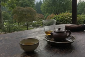 A contemplative and peaceful tea-drinking moment is set upon a weathered, dark wooden table overlooking a lush, verdant garden. The focus is on a traditional setup for tea service, featuring a small, rustic brown clay teapot resting on a matching shallow dish, a glass fair cup holding a small amount of golden-colored steeped tea, and an individual ceramic teacup filled with the warm brew. The scene conveys a sense of slow, deliberate ritual, framed by the blurred, natural greenery of the outdoors, which provides a calming and verdant backdrop to the simple yet elegant arrangement on the rustic table.