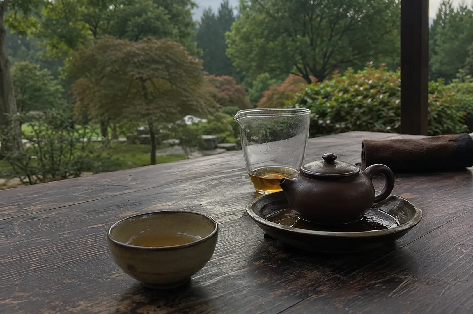 1 - Why Not Every Tea Deserves Your Attention A contemplative and peaceful tea-drinking moment is set upon a weathered, dark wooden table overlooking a lush, verdant garden. The focus is on a traditional setup for tea service, featuring a small, rustic brown clay teapot resting on a matching shallow dish, a glass fair cup holding a small amount of golden-colored steeped tea, and an individual ceramic teacup filled with the warm brew. The scene conveys a sense of slow, deliberate ritual, framed by the blurred, natural greenery of the outdoors, which provides a calming and verdant backdrop to the simple yet elegant arrangement on the rustic table.