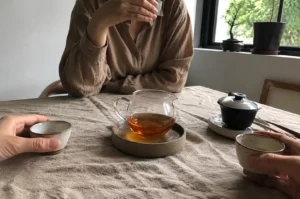 Two individuals are seated across from each other at a table covered in a textured, light beige cloth, sharing a tea service. A central glass pitcher filled with amber-colored tea sits on a rustic ceramic coaster, flanked by a black tea bowl with a lid on a saucer to the right and small, handmade-style ceramic cups held by the hands of each person. The person in the background wears a long-sleeved, earth-toned button-down shirt, and the setting is illuminated by soft natural light coming from a nearby window, where a small bonsai tree rests on the sill, creating a calm, minimalist atmosphere.