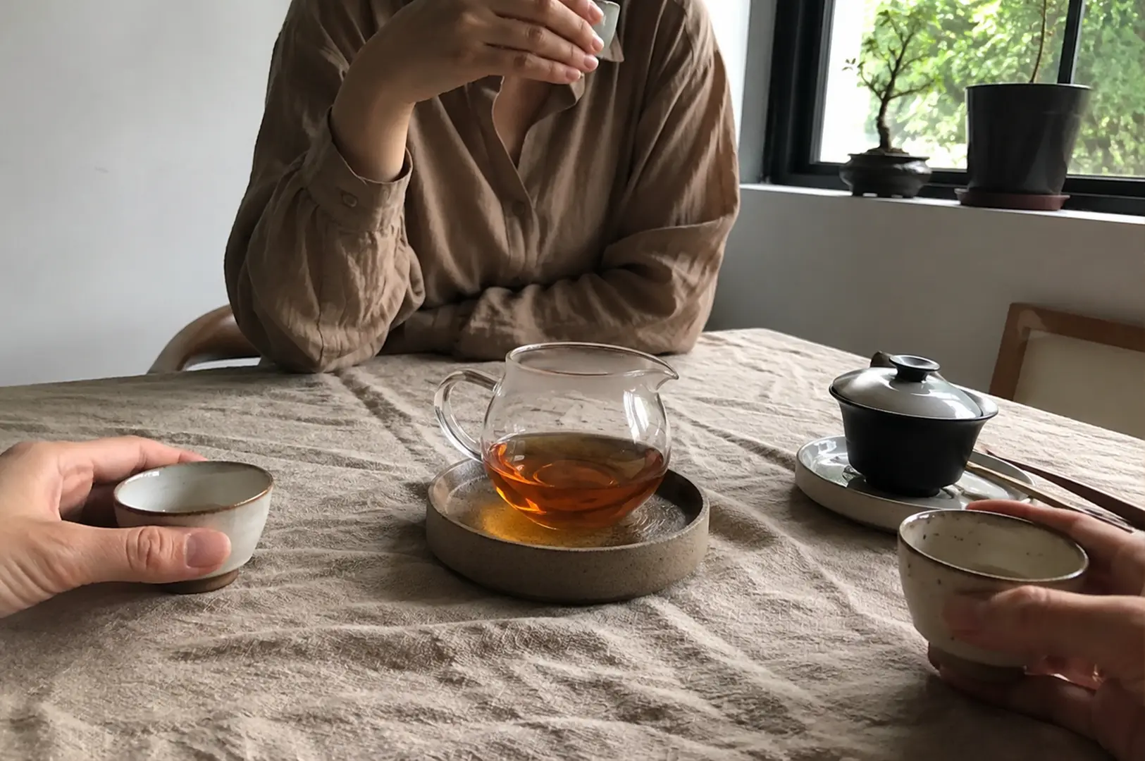 1 - Why Some Teas Taste Better When Shared Two individuals are seated across from each other at a table covered in a textured, light beige cloth, sharing a tea service. A central glass pitcher filled with amber-colored tea sits on a rustic ceramic coaster, flanked by a black tea bowl with a lid on a saucer to the right and small, handmade-style ceramic cups held by the hands of each person. The person in the background wears a long-sleeved, earth-toned button-down shirt, and the setting is illuminated by soft natural light coming from a nearby window, where a small bonsai tree rests on the sill, creating a calm, minimalist atmosphere.