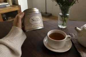 A person wearing a cozy, cream-colored ribbed knit sweater holds up a cylindrical silver tea tin over a dark wooden table. The tin features a decorative, light purple patterned label that reads "Black Tea", "No. 25", and "BELLOCQ" in elegant typography. On the table below, a matching speckled off-white ceramic teacup and saucer are filled with a warm, amber-colored tea, positioned next to a partially visible white ceramic teapot resting on a brown cloth napkin. In the soft-focused background, a ribbed glass vase holds a fresh bouquet of small white and pink wildflowers, adding a peaceful, serene ambiance to the warm indoor setting.