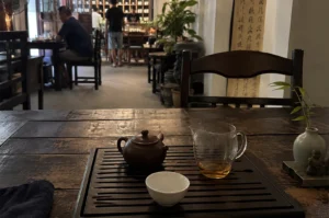 A cozy and authentic traditional Chinese teahouse unfolds from a first-person perspective at a dark wooden table. The foreground features a slatted Gongfu tea tray holding a small, matte brown clay teapot, a clear glass fairness cup partially filled with amber-colored tea, and an empty, pristine white porcelain teacup waiting to be filled. To the right of the tray, a small celadon-green vase cradles a delicate bamboo sprig next to a tiny stone Buddha figurine, while a dark tea cloth rests on the left. In the midground, an empty, dark-stained wooden chair stands in front of a vertical wall scroll adorned with elegant Chinese calligraphy, flanked by a vibrant green potted plant. The softly blurred background reveals the atmospheric depth of the shop, where a patron in a dark blue shirt sits at a distant table, and staff members stand near a massive, grid-like wooden shelving unit packed with traditional tea canisters under warm, moody lighting.