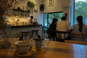 Captures the cozy, warm interior of a tea house or café from a low-angle perspective looking across a wooden table. In the immediate foreground, a glass teapot filled with light amber tea sits next to a rustic, textured ceramic teacup on a small wooden saucer, with a tall glass vase holding dried flowers visible on the left. The background reveals a warmly lit service counter where a barista in an apron is working, framed by shelves holding teapots, canisters, and hanging green plants. To the right, several patrons sit with their backs to the camera at wooden tables, facing a large floor-to-ceiling window that looks out onto lush, green outdoor trees, allowing soft daylight to mix with the café's golden, ambient indoor lighting.