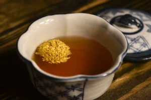 A close-up shot of an elegant white ceramic teacup with blue grid-like patterns on its base, resting on a dark rustic wooden table. A single, large golden-yellow chrysanthemum flower blooms beautifully as it floats on the surface of the amber-colored herbal tea inside the cup. In the blurred background, the matching patterned lid of the teacup lies slightly askew on the wooden surface.