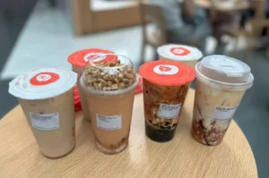 Six varied bubble tea drinks from LiHO Tea are arranged on a light wood tabletop, featuring iced milk teas with red sealed lids, a Thai tea smoothie crowned with toasted cereal puffs under a clear dome lid, and brown sugar pearl milk teas displaying rich syrup marbling through milk and tapioca pearls.