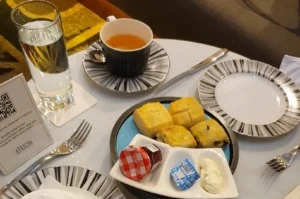 A close-up view of a high tea setting on a white table features a striped teacup and saucer paired with a plate of four golden-brown scones, served alongside a small dish containing a miniature jar of strawberry jam, a butter portion, and a dollop of clotted cream. A tall glass of water and a menu stand with a QR code are also visible on the table, which is set with a silver knife and fork, highlighting the refined details of the afternoon tea service.