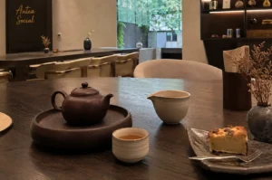 A dark, wood-grain tea table setup in a serene and minimalist teahouse interior, featuring a traditional brown Yixing clay teapot sitting on a matching round tray, a small teacup filled with clear amber tea, a light ceramic fairness pitcher, and a slice of golden cake served on a rustic dark plate alongside a small vase of dried flowers. In the background, soft ambient lighting illuminates neutral-toned textured walls, modern low-backed seating, elegant dark display shelving with teaware, and the cursive "Antea Social" brand signage, with a large window opening toward lush outdoor greenery.