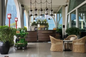 An elegant outdoor covered corridor at The Clifford Pier featuring high arched ceilings with industrial hanging lantern chandeliers, dark wood service bars bearing "THE CLIFFORD PIER" sign, a green drink cart, and woven wicker armchairs arranged around a small coffee table.