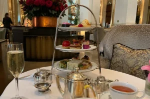 A three-tiered silver stand filled with delicate afternoon tea delicacies sits centered on a round table draped with a white tablecloth, featuring finger sandwiches on the bottom tier, colorful bite-sized pastries and a glossy red apple-shaped dessert in the middle, and fresh scones with jam on top. Arranged on the table around the stand are silver teapots, a silver tea strainer, a porcelain cup filled with amber tea, a tall flute of sparkling champagne, and a pink flower in a vase, all set against the opulent backdrop of an grand hotel lounge with plush armchairs and a massive red floral centerpiece.