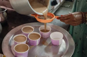 A person’s hand is seen straining hot, milky masala chai through a small orange plastic sieve from a large metal pot into several paper cups arranged on a metallic tray, illustrating the preparation process in a busy kitchen setting.
