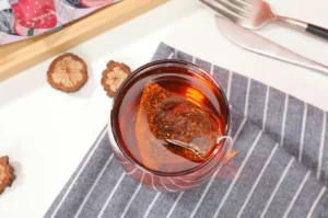 A top-down view shows a clear glass filled with rich reddish-amber tea steeping from a mesh pyramid teabag, resting on a grey and white striped fabric napkin. Dried fruit slices are scattered nearby on the clean white surface, while a wooden tray edge and silver cutlery frame the upper corner of the scene.