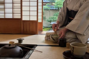 A woman dressed in a soft-toned kimono kneels on a tatami mat in a traditional Japanese tea room, carefully whisking matcha in a rustic ceramic bowl with a bamboo chasen. Before her on the mat are various tools of the tea ceremony, including an iron kettle, a bamboo ladle, and a small tea caddy, all arranged with precision. In the background, open shoji sliding doors frame a serene view of a lush green garden, emphasizing the peaceful and focused atmosphere of the ceremony captured.