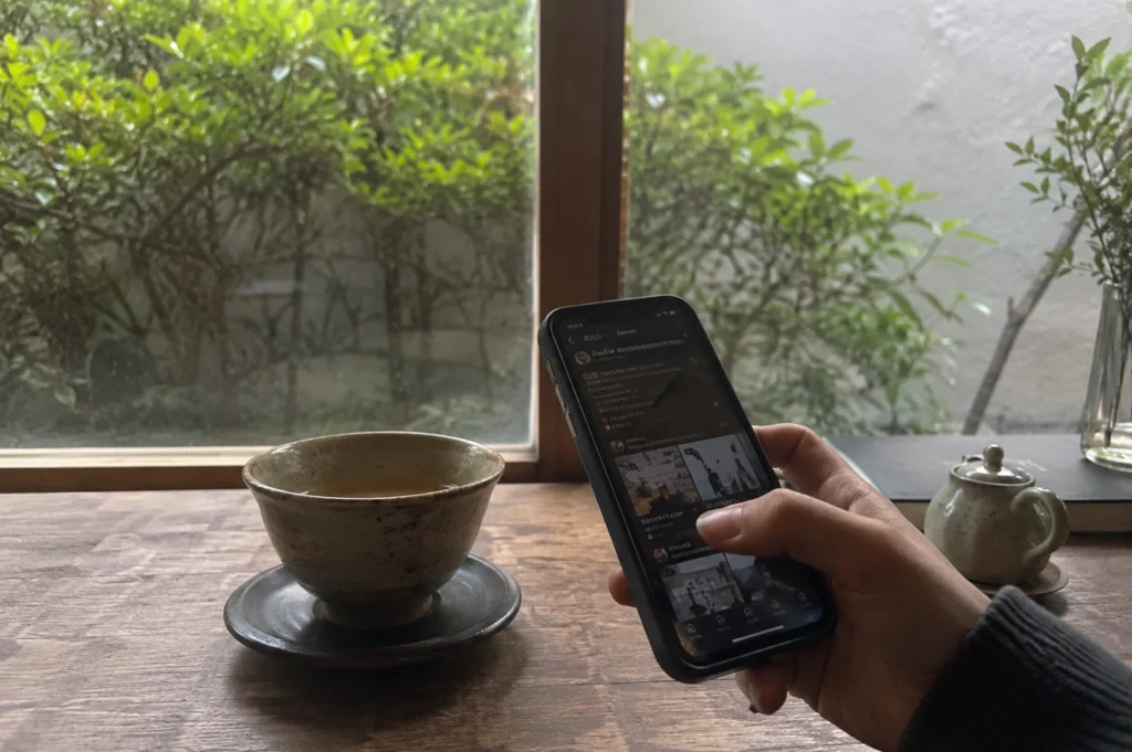 A hand holding a dark-cased smartphone dominates the foreground of this serene, eye-level shot, with a thumb actively scrolling through a social media or content feed on the screen. To the left of the phone, a rustic, speckled ceramic teacup sits on a matching saucer on a worn wooden table, suggesting a quiet moment of relaxation. Through a large, rectangular window in the background, a lush green garden with dense foliage provides a soft-focus, natural backdrop that contrasts with the digital activity on the phone. Completing the calm, minimalist table setting are a small ceramic teapot and a glass vase with greenery, all captured in a way that emphasizes a peaceful, modern lifestyle balancing digital engagement with contemplative downtime.