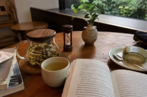 A cozy, serene table setup for reading and tea features an open book lying flat in the foreground, flanked by a rustic ceramic cup filled with pale green tea and a stack of paperbacks on the left. On the warm wooden tabletop behind them, a clear glass teapot with a wooden lid steeps loose-leaf tea on a cork coaster, accompanied by a small wooden hourglass timer, a small ceramic vase holding a green leafy sprig, and a plate with a matcha swiss roll dessert served with a spoon.