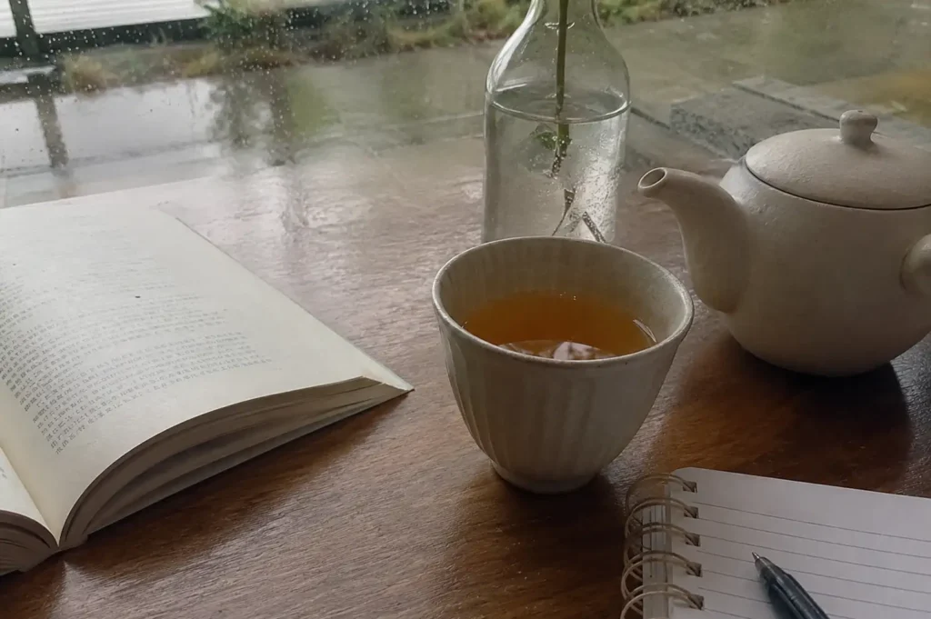 A cozy, reflective scene unfolds on a smooth wooden table positioned in front of a window beaded with raindrops from a rainy day outside. On the left, an open book with printed text rests flat on the surface, catching soft light. In the center, a handleless ceramic teacup with vertical ridges holds a warm, golden tea, flanked by an off-white ceramic teapot to the right and a clear glass bottle with a single flower stem in water behind it. In the bottom right foreground, a spiral-bound notebook lies open next to a black pen, completing the quiet reading and writing setup.