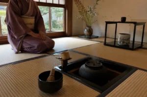 A wide-angle shot of a traditional Japanese tea room with a practitioner in a muted purple kimono sitting in the seiza posture next to a large window overlooking a green garden. In the foreground, a square sunken hearth built into the tatami floor holds a dark iron kettle with faint steam rising from it. A black bowl containing a bamboo whisk sits to the left of the hearth, while the right side of the room features a black tiered display stand with a decorated ceramic jar and a tall, rustic vase holding a minimalist arrangement of wild branches.