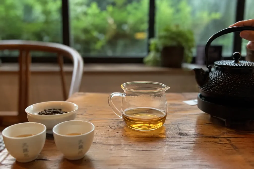 Set on a warm, rustic wooden table in front of a window overlooking vibrant green foliage, a traditional tea set includes a clear glass pitcher filled with amber-colored tea, a black textured cast-iron teapot being held by its handle, a small white bowl with loose tea leaves, and two white ceramic teacups featuring Chinese characters printed on their sides.