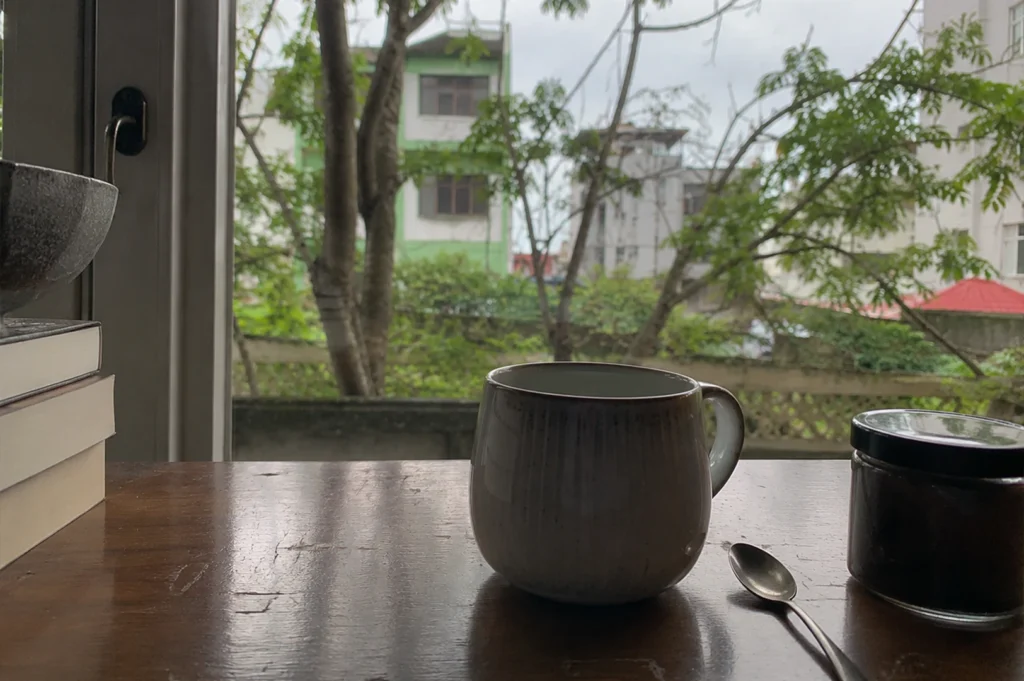 The image captures a quiet, contemplative scene set on a dark, polished wooden surface near a window. A rounded, light-toned ceramic mug sits prominently in the center, accompanied by a small silver spoon and a glass jar with a black lid nearby. To the left, a stack of books provides a grounding element, while through the window, a lush view of trees and soft-hued urban buildings fills the background, creating a serene and cozy atmosphere under a diffused, overcast sky.