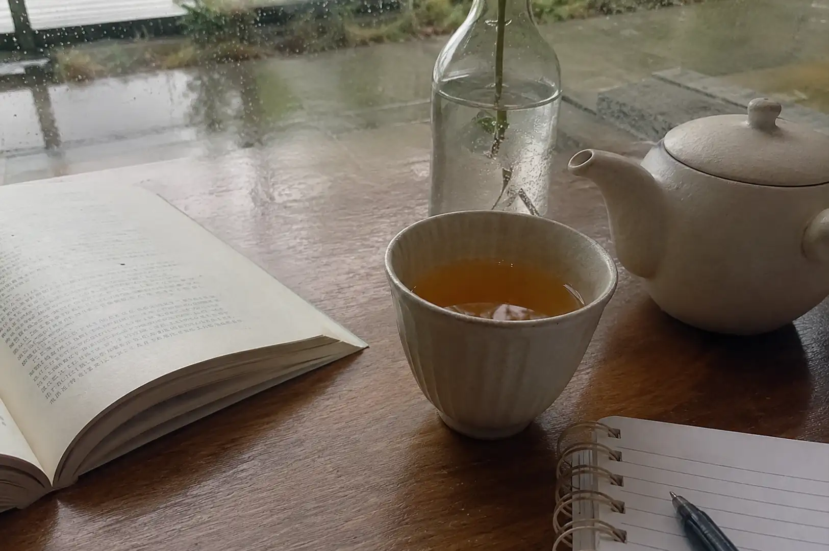 1 - To the Tea Lovers and the Tea That Holds Us A cozy, reflective scene unfolds on a smooth wooden table positioned in front of a window beaded with raindrops from a rainy day outside. On the left, an open book with printed text rests flat on the surface, catching soft natural light. In the center, a handleless ceramic teacup with fine vertical ridges holds a warm, golden-amber tea, flanked by a matching off-white ceramic teapot on the right and a tall, clear glass bottle with a single plant stem in water directly behind it. In the bottom right foreground, a spiral-bound notebook lies open beside a black pen, completing the quiet study and reading setup.