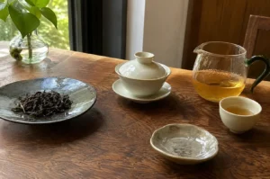 Arranged neatly on a rich wooden tabletop, a traditional Gongfu tea setup includes a shallow dark grey ceramic plate holding a pile of loose dark tea leaves, a white covered gaiwan on a saucer, a small white tasting cup filled with golden tea, a small rustic saucer, and a clear glass pitcher with a green handle containing warm amber tea, with a small potted plant sitting near a bright window in the background.