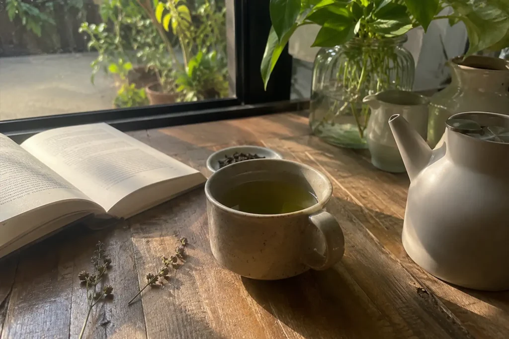 A peaceful, sunlit morning scene is set on a rustic wooden table positioned next to a large window that looks out onto a lush, green outdoor garden. An open book with dense text lies flat on the left side of the table, catching warm sunlight across its pages. In the center, a speckled ceramic mug filled with a pale green tea is flanked by two delicate, dried floral stems resting on the wood. To the right, a modern white teapot and a small ceramic creamer pitcher sit near a large glass vase holding vibrant leafy green plant cuttings.