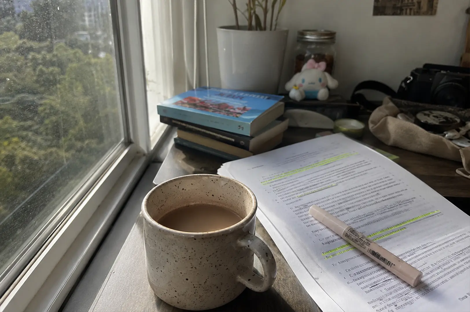 1 - Why We Keep Romanticising Tea Moments That Never Happened A tranquil study scene is set beside a window looking out onto lush green trees, with a speckled ceramic mug filled with coffee taking center stage. To the right, a stack of printed documents with highlighted text lies on the desk, accompanied by a light-colored highlighter pen. The background features a small potted plant, a glass jar, a plush character toy, and a camera, all arranged on a desk that catches the soft natural light streaming through the window.