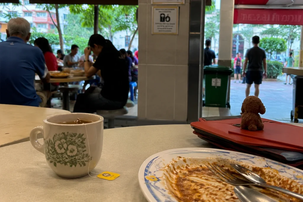 The image captures a casual, intimate scene at an open-air food center in Singapore. In the foreground, a white ceramic mug with a delicate green floral pattern holds hot tea, with its yellow tea bag tag resting on a light-colored table next to a plate featuring remnants of a meal and stainless steel cutlery. Perched atop a red leather-bound notebook lying nearby is a small, brown plush toy resembling a miniature poodle. The background, which is slightly out of focus, reveals a bustling, partially shaded hawker center filled with various diners sitting at tables. A vertical pillar prominently displays signs for a "Tray Return Point," while in the distance, people walk across a paved path beneath lush green trees, suggesting a warm and relaxed daytime atmosphere.