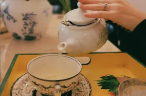 A close-up shot capturing a person’s hand pouring steaming hot tea from a white patterned ceramic teapot into a matching delicate teacup with floral details, which sits on an intricate saucer atop a yellow wooden tray.