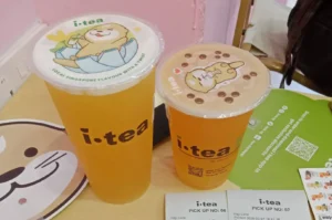 Two plastic cups from iTea filled with vibrant orange tea beverages resting on a light wooden table alongside receipt slips. The cup on the left is tall with a white sealed plastic lid featuring a cute otter holding a drink, while the slightly shorter cup on the right displays a beige-tinted sealed lid with an otter resting surrounded by tapioca pearls.