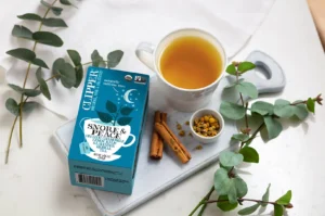 A cozy flat-lay arrangement featuring a teal box of Clipper "Snore & Peace" Organic Chamomile, Lemon Balm & Lavender Herbal Tea on a white wooden cutting board. Beside the box, a filled white teacup sits next to whole cinnamon sticks, a small white bowl of dried chamomile flowers, and lush green eucalyptus branches spread across a marble surface.