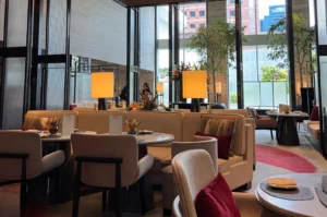 A wide shot reveals a bright, upscale hotel lounge featuring plush beige curved booths and light grey upholstered armchairs arranged around round marble tables. Warm rectangular floor lamps with fabric shades cast a cozy glow, while floor-to-ceiling glass windows show indoor potted trees and city buildings in the background.