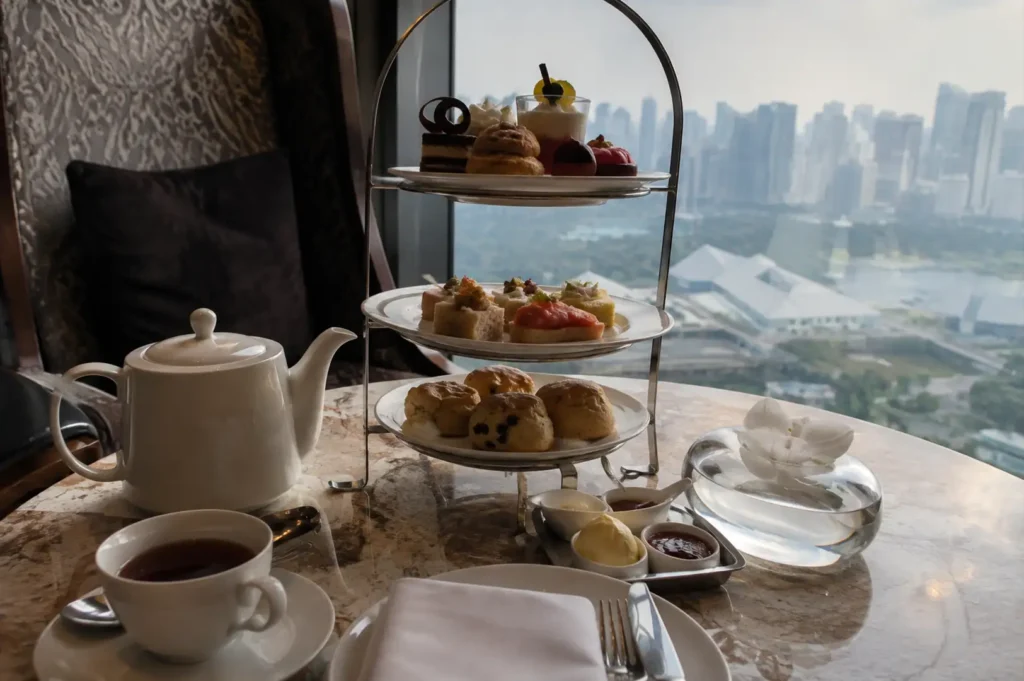 A eye-level table arrangement of an elegant afternoon high tea service set on a polished marble table next to a high-rise window overlooking a hazy city skyline, featuring a silver three-tiered stand loaded with delicate miniature desserts on top, savory finger sandwiches in the middle, and freshly baked scones on the bottom, accompanied by a white porcelain teapot, a filled teacup on a saucer, a place setting with folded cloth napkin and silver cutlery, a small metal tray with condiment bowls of clotted cream and jam, and a clear glass bowl containing a white orchid stem.