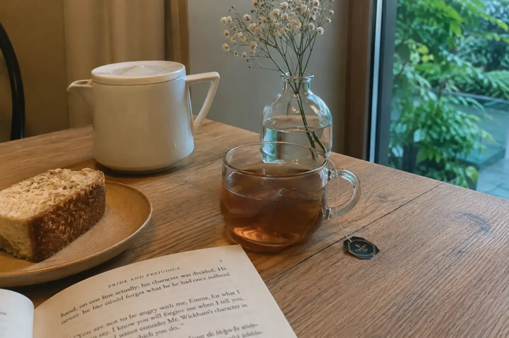 A warm, high-angle shot of a serene cafe moment set on a wooden table near a glass window revealing lush outdoor greenery, featuring an open copy of Pride and Prejudice in the foreground next to a glass mug of steeped tea with its tag hanging over the side, accompanied by a slice of toasted bread or loaf cake on a neutral-toned ceramic plate, a sleek white ceramic teapot, and a clear glass vase holding delicate white baby's breath blossoms.